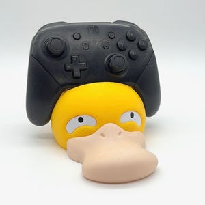 May include: A black Nintendo Switch Pro controller rests on a yellow and tan Psyduck-shaped stand. The stand features the character's distinctive beak and eyes, creating a fun and functional accessory for gamers.