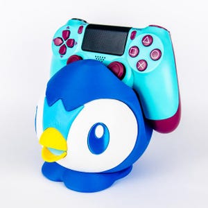 May include: A blue and white Piplup-themed controller holder with a teal and magenta game controller resting on top. The controller has a black center panel and magenta buttons.