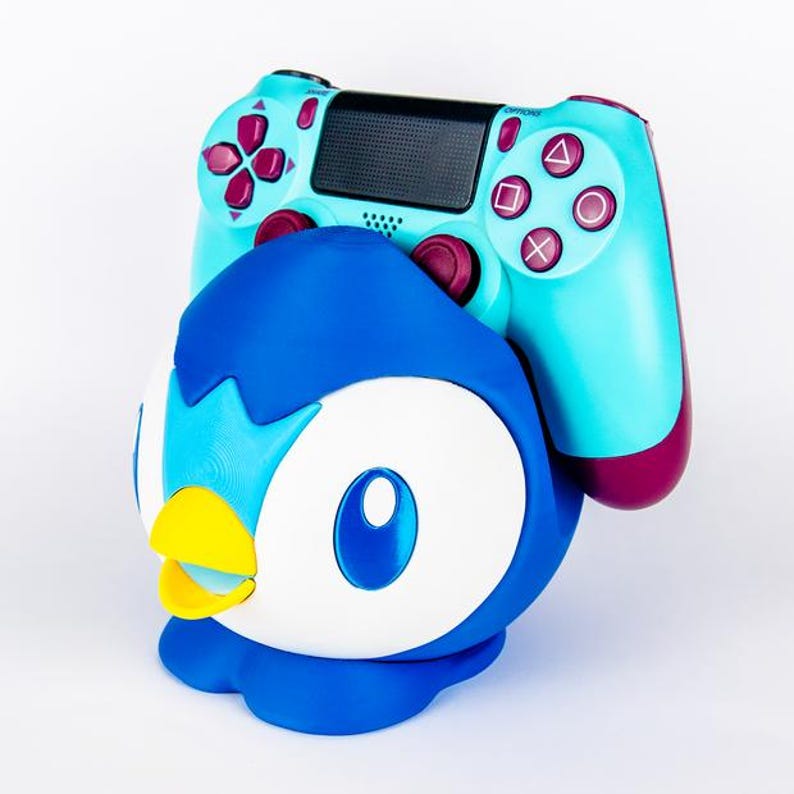 May include: A blue and white Piplup-themed controller holder with a teal and magenta game controller resting on top. The controller has a black center panel and magenta buttons.