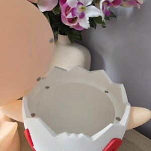 May include: A white, egg-shaped decorative container with red and blue accents. The container has a circular opening and is designed with a cartoon aesthetic. The background includes a bouquet of pink and purple flowers.