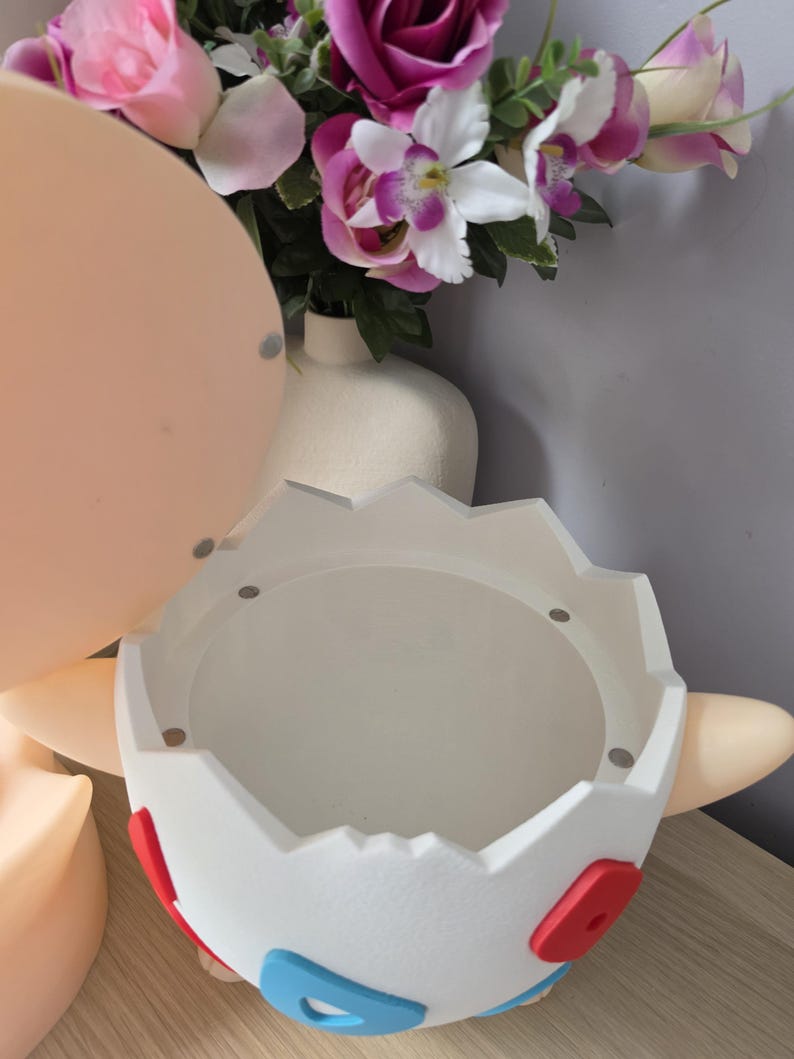 May include: A white, egg-shaped decorative container with red and blue accents. The container has a circular opening and is designed with a cartoon aesthetic. The background includes a bouquet of pink and purple flowers.