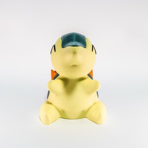 May include: A yellow figurine of a cartoon character with a blue head, black eyes, and orange accents. The figurine is sitting with its arms and legs spread out. The background is plain white.
