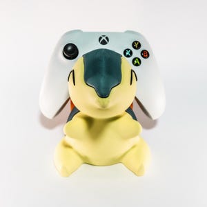 Peut inclure: Une figurine jaune et sarcelle d'un personnage de dessin animé est placée devant une manette Xbox blanche. La manette a des boutons noirs et des lettres colorées. La figurine a des lignes noires et des accents orange.