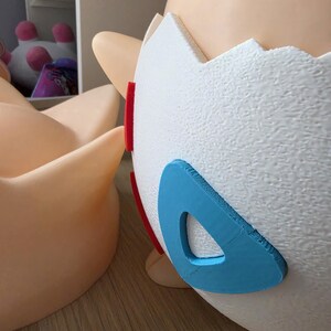 May include: A close-up of a Togepi figurine, a Pok&eacute;mon character, with a white eggshell body and tan skin. The eggshell has red and blue accents. The figurine is on a light brown surface.