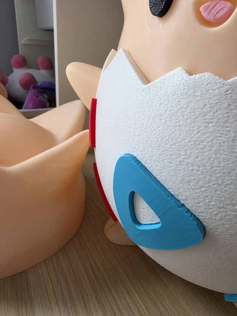 May include: A close-up of a Togepi figurine, a Pok&eacute;mon character, with a white eggshell body and tan skin. The eggshell has red and blue accents. The figurine is on a light brown surface.