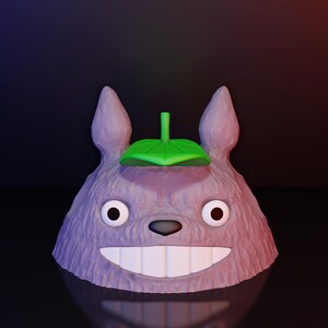 Może przedstawiać: Rendowana w 3D figurka przypominająca Totoro. Figurka jest gł&oacute;wnie lawendowa z teksturowaną powierzchnią, dużymi białymi zębami i zielonym liściem na głowie. Oczy są czarno-białe, na ciemnym tle.