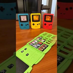 May include: A collection of colorful handheld gaming consoles in blue, yellow, and red, with a green cartridge holder. The consoles have a classic design with buttons and a screen. The cartridge holder is open, displaying game cartridges.