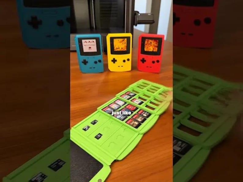 May include: A collection of colorful handheld gaming consoles in blue, yellow, and red, with a green cartridge holder. The consoles have a classic design with buttons and a screen. The cartridge holder is open, displaying game cartridges.