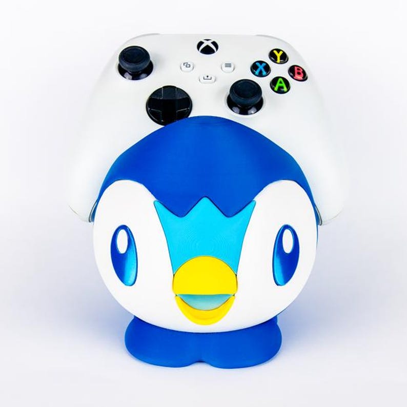 May include: A white Xbox controller rests atop a blue, white, and yellow Piplup figure. The controller has black joysticks and colored buttons. The Piplup figure has large blue eyes and a yellow beak. The overall theme is gaming.