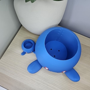 May include: Two blue ceramic planters shaped like cartoon characters. The larger planter has a round body with two arms and legs. The smaller planter has a round body with two arms. Both planters have a hole in the top for planting.
