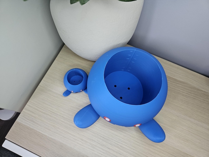 May include: Two blue ceramic planters shaped like cartoon characters. The larger planter has a round body with two arms and legs. The smaller planter has a round body with two arms. Both planters have a hole in the top for planting.