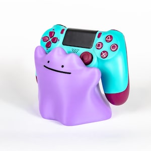 Op de afbeelding: Een paarse Ditto-vormige controllerhouder houdt een turquoise en bordeauxrode gamecontroller vast. De controller heeft bordeauxrode knoppen en een zwart middenpaneel. De houder heeft een eenvoudige zwarte glimlach.