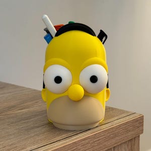 May include: A yellow pen holder shaped like Homer Simpson's head, with large white eyes, a yellow nose, and a tan mouth. The pen holder has a black band on top and holds several pens and markers.