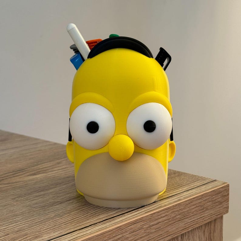 May include: A yellow pen holder shaped like Homer Simpson's head, with large white eyes, a yellow nose, and a tan mouth. The pen holder has a black band on top and holds several pens and markers.