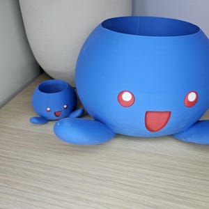 May include: Two blue ceramic planters shaped like a cartoon octopus with red eyes and a mouth. The larger planter is in the foreground and the smaller planter is in the background.