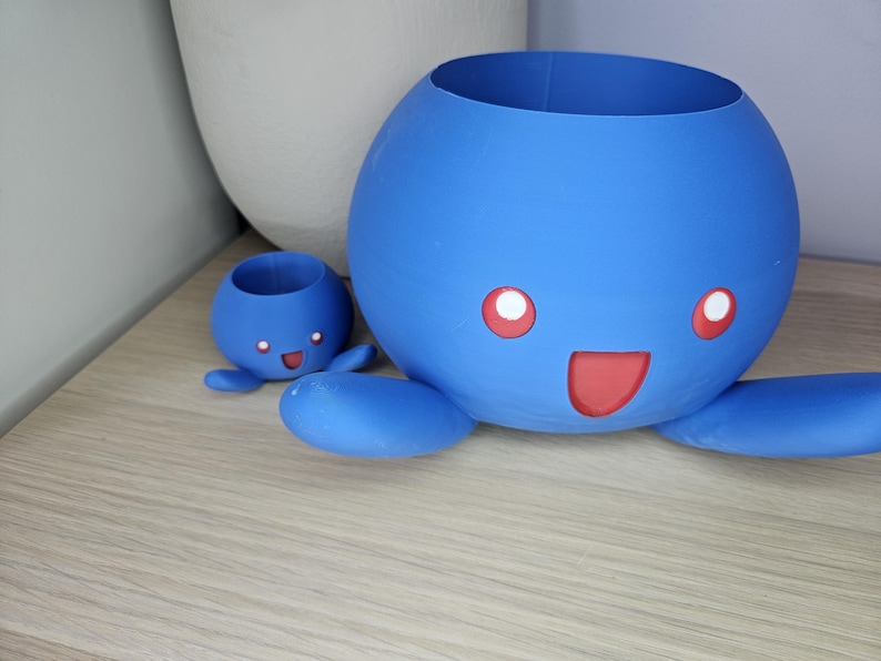 May include: Two blue ceramic planters shaped like a cartoon octopus with red eyes and a mouth. The larger planter is in the foreground and the smaller planter is in the background.