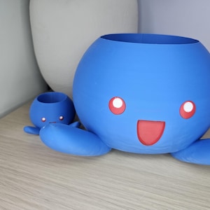 May include: Two blue ceramic octopus planters with red smiling faces. The larger planter has a wider opening and the smaller planter has a narrower opening.