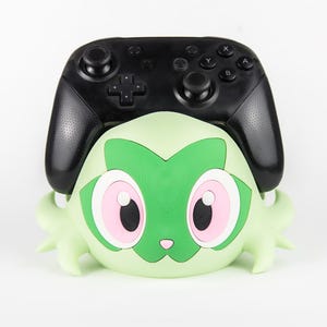 Puede incluir: Un mando de videojuegos inalámbrico negro apoyado sobre un soporte de color verde claro con forma de personaje. El soporte presenta una cara estilizada con grandes ojos rosas, una pequeña nariz rosa y marcas verde oscuro para sujetar el mando.