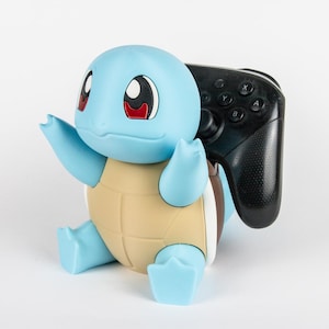 Può includere: Una statuetta del personaggio Squirtle in azzurro e beige che funge da supporto per controller. La figura ha grandi occhi con pupille rosse e tiene un controller per videogiochi nero con pulsanti etichettati A, B, X e Y nel braccio destro.