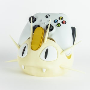 May include: A cream-colored, cartoon-style controller holder shaped like a character's head, with a gold accent and white details. A white Xbox controller rests in the holder. The controller has black buttons and colorful action buttons.