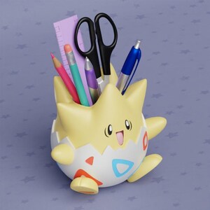 May include: A yellow and white character-shaped desk organizer holding school supplies. The organizer has a smiling face and holds a pink pencil, a teal pencil, a purple pen, black scissors, a blue pen, and a purple ruler.