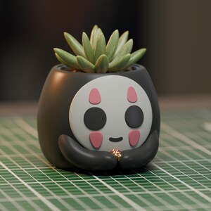 May include: A black and white ceramic planter shaped like a character from the movie Spirited Away. The planter has a green succulent plant growing inside.