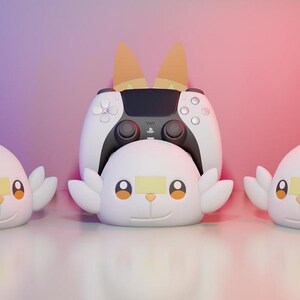 Könnte beinhalten: Drei weiße Controller-Ständer im Charakter-Design mit großen orangefarbenen und gelben Ohren, kleinen Flügeln und einem lächelnden Gesicht. Der mittlere Ständer hält einen schwarz-weißen kabellosen Gamecontroller mit PlayStation-Logo.