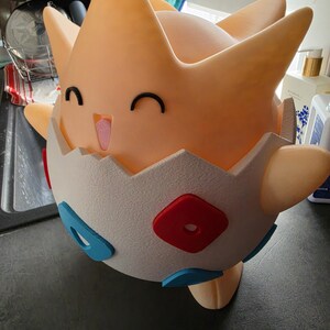 May include: A 3D-printed Togepi figure in a cheerful pose. The figure is mainly peach-coloured with a white eggshell base. It features black eyes, a pink tongue, and red and blue accents on the eggshell.