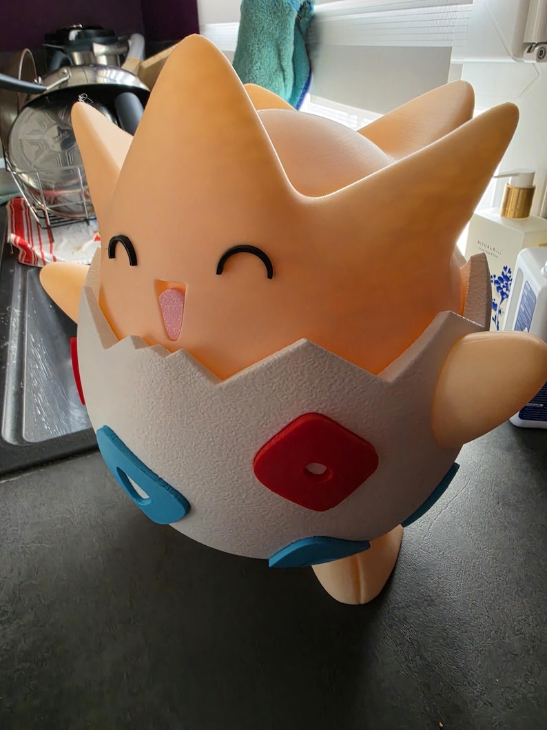 May include: A 3D-printed Togepi figure in a cheerful pose. The figure is mainly peach-coloured with a white eggshell base. It features black eyes, a pink tongue, and red and blue accents on the eggshell.