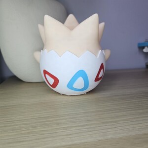 May include: A Togepi figurine, a Pok&eacute;mon character, is displayed on a wooden surface. The figurine is white with red and blue accents, and a cream-colored head. The background includes a white vase and a blue object.