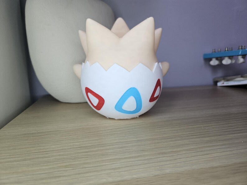 May include: A Togepi figurine, a Pok&eacute;mon character, is displayed on a wooden surface. The figurine is white with red and blue accents, and a cream-colored head. The background includes a white vase and a blue object.
