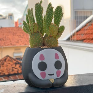 May include: A black and white No Face character planter with a pink smile and eyes. A green cactus plant is growing in the planter.