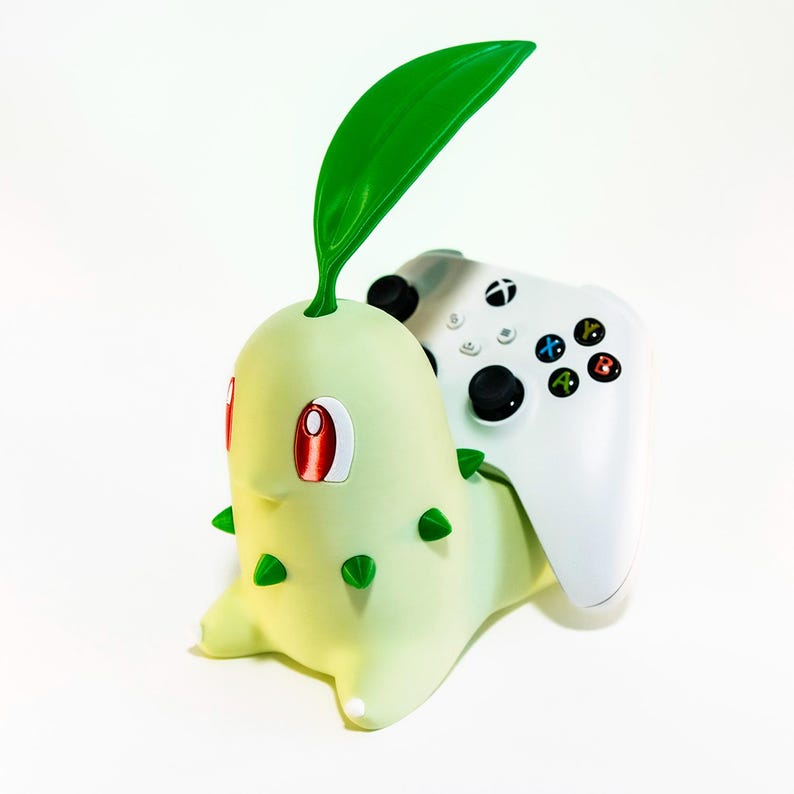 May include: A light green Chikorita-shaped controller holder with a white Xbox controller. The holder has red eyes, green spikes, and a large green leaf. The controller has black buttons and coloured action buttons.