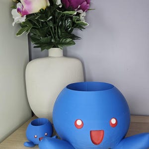 Op de afbeelding: Twee blauwe keramische potten in de vorm van een cartoon octopus met rode glimlachende gezichten. De grotere pot heeft een witte vaas met roze en paarse bloemen erachter.