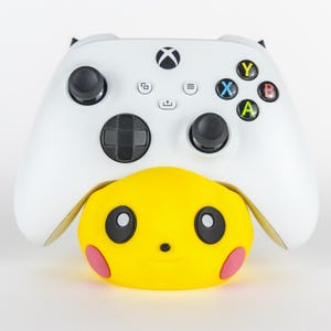 Pu&ograve; includere: Un controller Xbox bianco con joystick e pulsanti neri poggia su un accessorio giallo a forma di Pikachu. L'accessorio ha occhi neri e cerchi rosa sulle guance. Il controller ha pulsanti colorati: verde, blu, rosso e giallo.