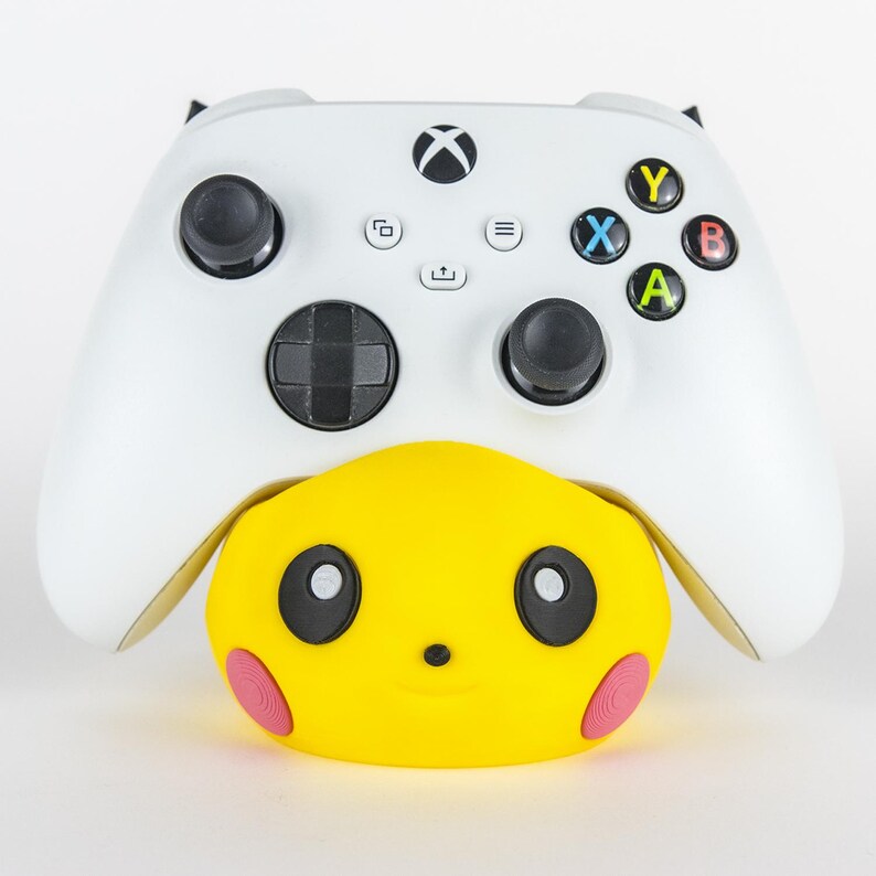 Pu&ograve; includere: Un controller Xbox bianco con joystick e pulsanti neri poggia su un accessorio giallo a forma di Pikachu. L'accessorio ha occhi neri e cerchi rosa sulle guance. Il controller ha pulsanti colorati: verde, blu, rosso e giallo.