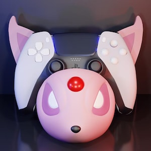 May include: A custom-designed PlayStation 5 controller with a pink and white color scheme, featuring a stylized character design. The controller has a pink face, white ears, and white controller pads. A red sphere is on the face.