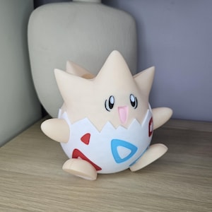 May include: A Togepi figurine, a Pokémon character, is depicted. The figure is primarily beige with white, red, and blue accents. It has a star-shaped head, small arms, and is posed on a wooden surface. The figurine is a collectible item.
