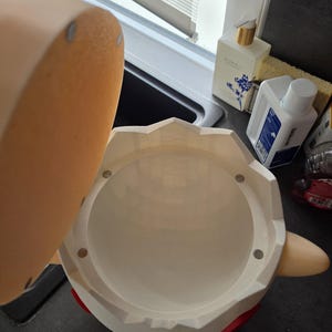 May include: A white container with a lid, featuring red and blue accents. The lid is a light tan colour. The container has a round opening and a cylindrical interior. The container is on a dark surface.