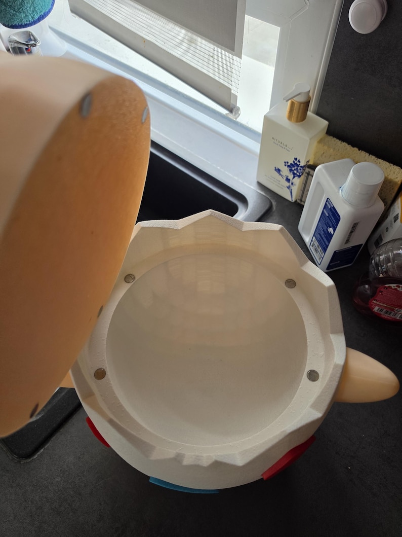May include: A white container with a lid, featuring red and blue accents. The lid is a light tan colour. The container has a round opening and a cylindrical interior. The container is on a dark surface.