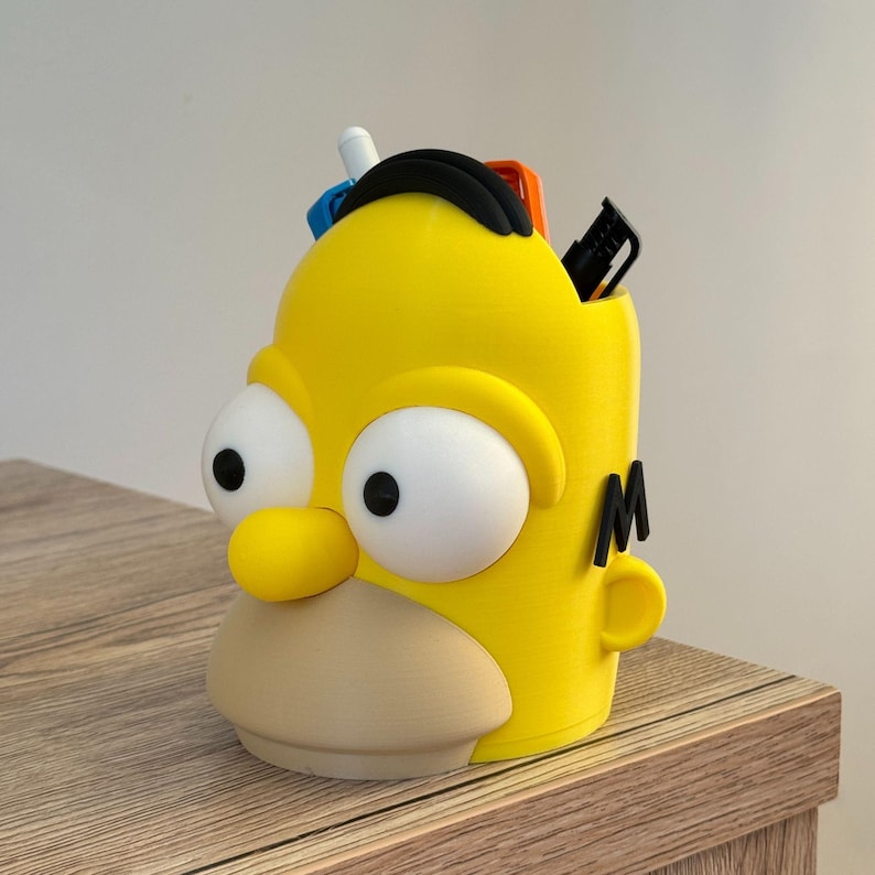 May include: A yellow pen holder shaped like Homer Simpson's head, with large white eyes and a beige mouth. The pen holder has black hair and the letter "M" on the side. Several pens and markers are visible inside the holder.