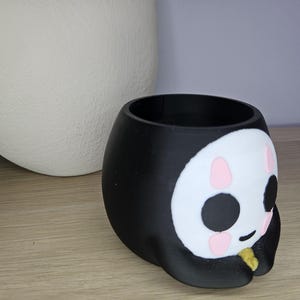 May include: Black and white ceramic planter shaped like a cartoon face with pink cheeks and a gold accent. The planter has a round opening at the top for planting.