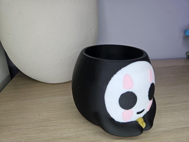May include: Black and white ceramic planter shaped like a cartoon face with pink cheeks and a gold accent. The planter has a round opening at the top for planting.