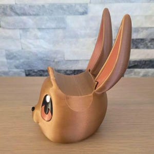 May include: A brown, 3D-printed bunny-shaped phone holder. The holder features large, upright ears with a peach-colored interior, and large eyes with a black pupil and a peach-colored iris. The holder is designed to cradle a phone.