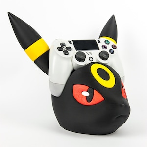 Peut inclure: Un support de manette Umbreon noir et jaune avec une manette PlayStation 4 blanche posée dessus. Le support présente des yeux rouges et un motif d'anneau jaune. La manette a des boutons noirs.