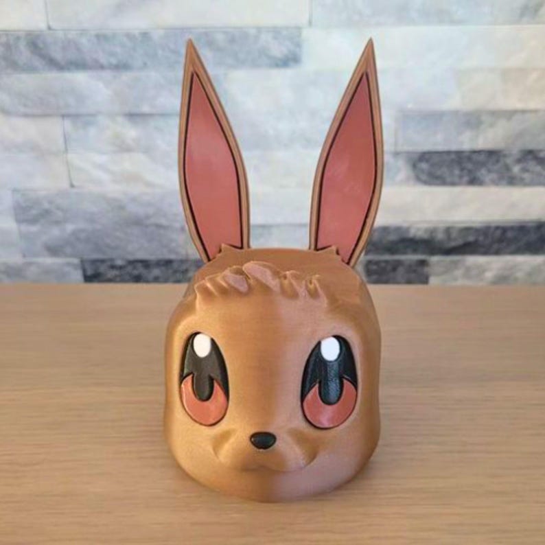 May include: A brown 3D-printed planter shaped like a cartoon bunny with large eyes and long ears. The ears have a pink interior and a brown outline. The planter is sitting on a wooden surface.