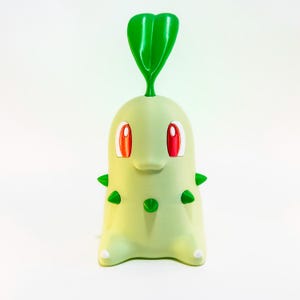May include: A light green Chikorita figure with red eyes and green spikes. The figure has a large green leaf on top and small white feet. The figure is a collectible item from the popular Pok&eacute;mon franchise.