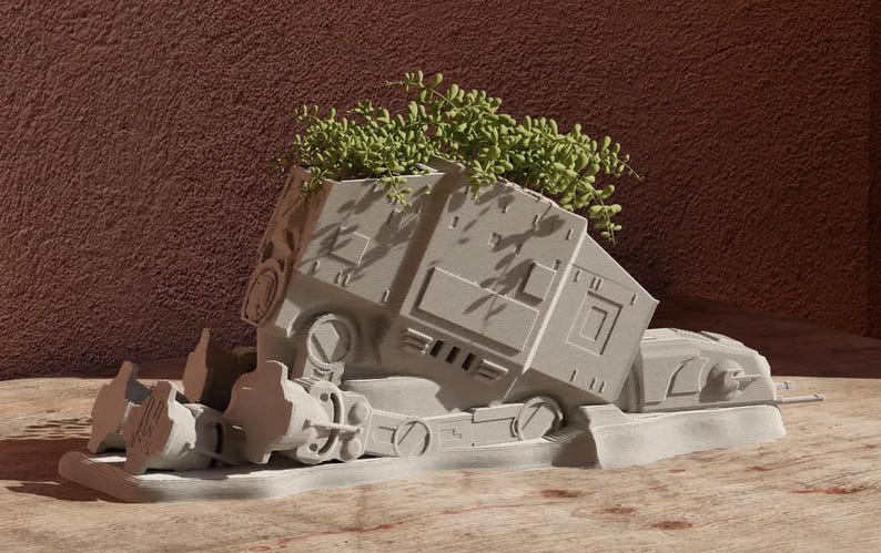 May include: A white ceramic planter shaped like a Star Wars AT-ST walker with green plants growing out of the top.