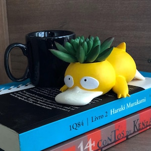 May include: A yellow duck-shaped planter with a green succulent plant inside. The planter is lying on its side on top of two books. The top book is blue and has the text "1Q84 | Livro 2 Haruki Murakami" printed on the spine. The bottom book is red and has the text "14 Contos de Kenza" printed on the spine.