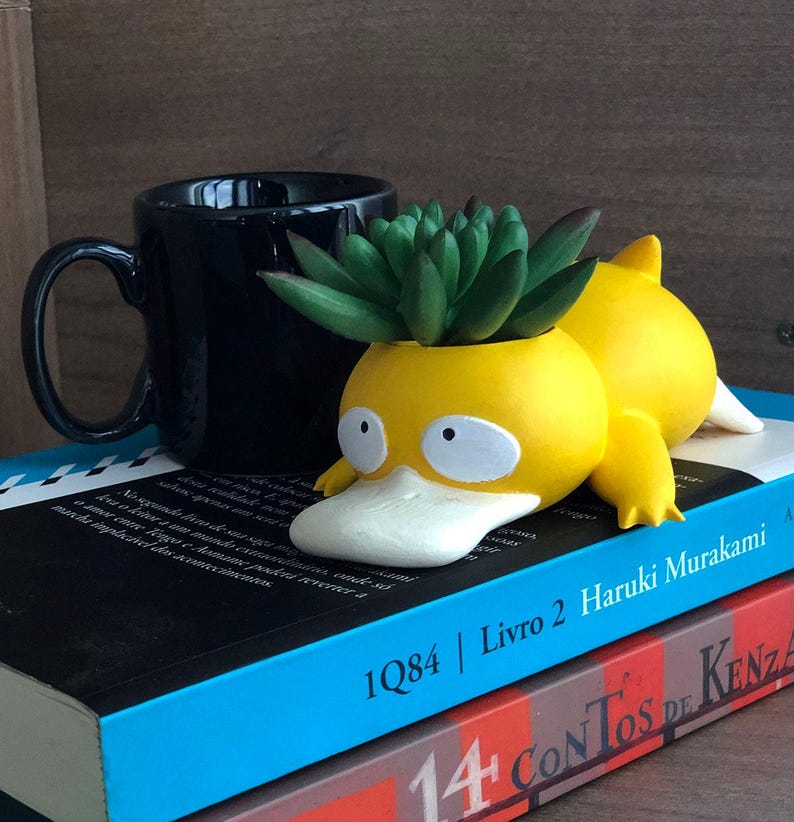 May include: A yellow duck-shaped planter with a green succulent plant inside. The planter is lying on its side on top of two books. The top book is blue and has the text "1Q84 | Livro 2 Haruki Murakami" printed on the spine. The bottom book is red and has the text "14 Contos de Kenza" printed on the spine.